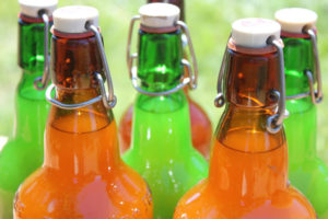 Close-up of six glass bottles with swing-top caps. Three are filled with an orange liquid and three with a green liquid. The bottles are arranged closely together, with sunlight reflecting off their surfaces.