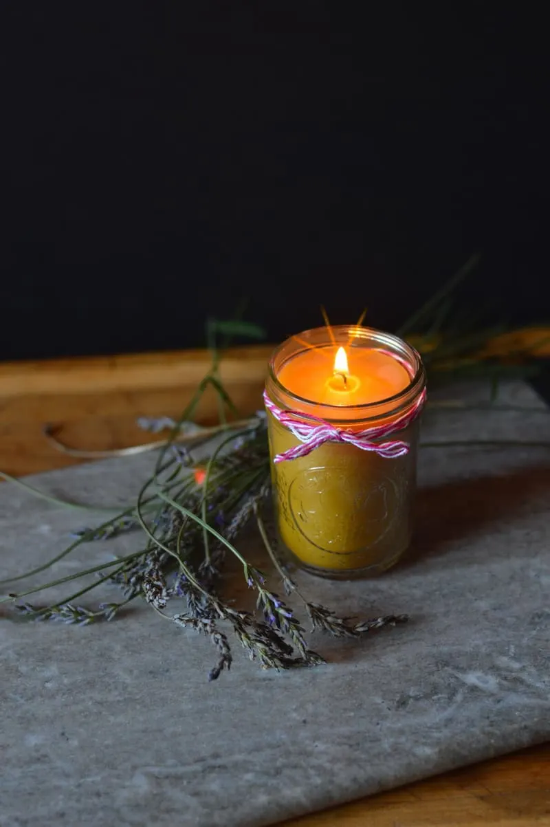 A lit beeswax candle in a mason jar sitting on a rock surrounded by sprigs of lavender.