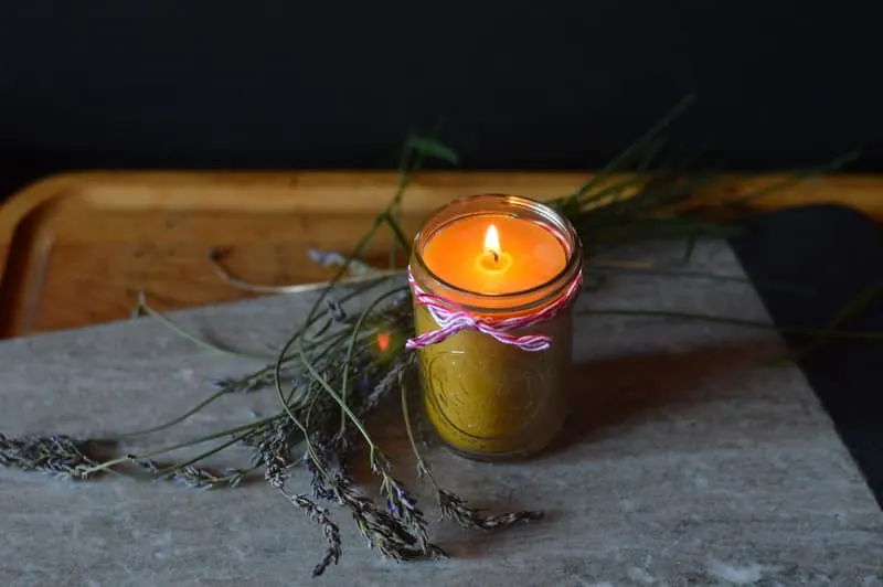 A lit beeswax candle in a mason jar sitting on a rock surrounded by sprigs of lavender.