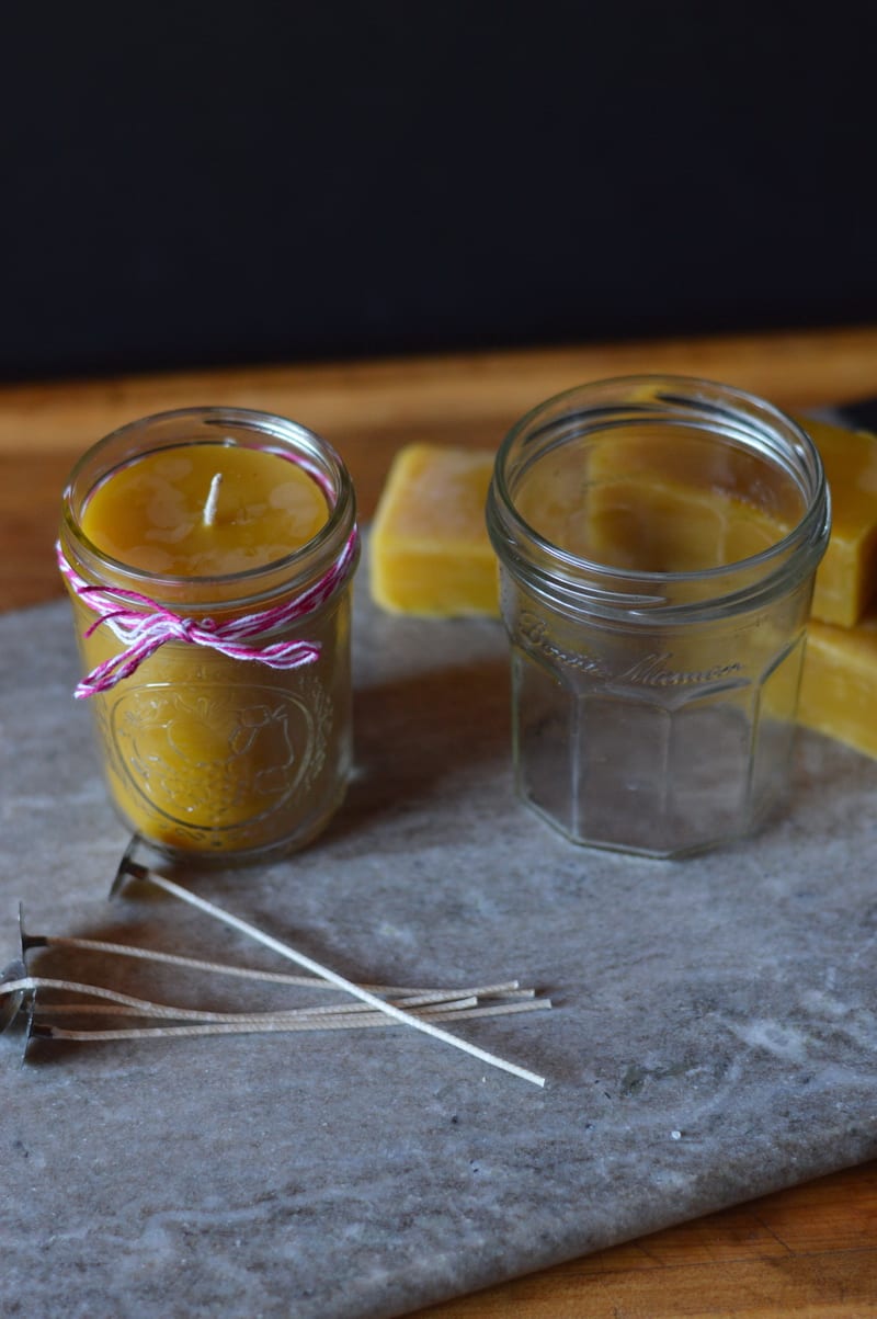 A beeswax candle in a jar sitting on a rock surrounded by an empty jar, candle wicks, and blocks of beeswax.