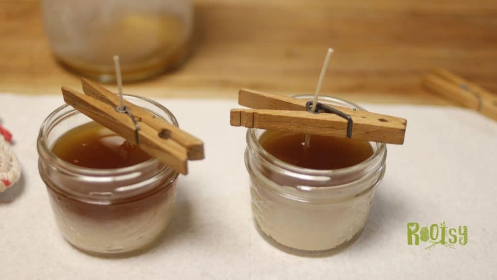 Two small glass jars filled with brown liquid and white wax, each with a wick held upright by a wooden clothespin. These charming beeswax candles sit on a white surface with a "Rootsy" logo in the bottom right corner.