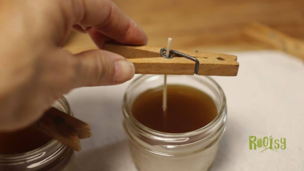 A hand holding a clothespin over a glass jar with liquid, preparing to make beeswax candles.