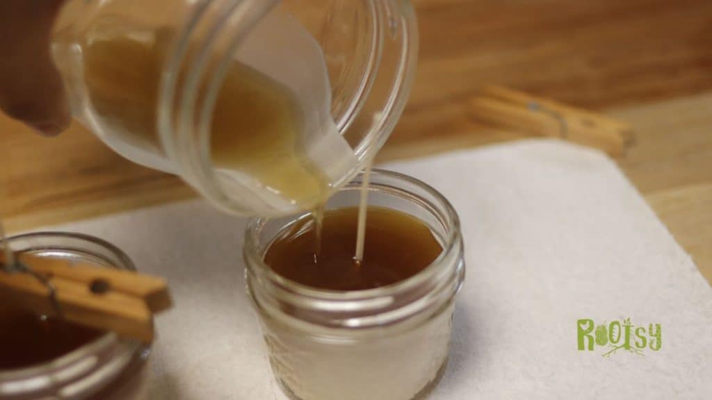 A hand pours melted beeswax from a glass jar into a small glass container of amber liquid, crafting beeswax candles on a white cloth. Wooden clothespins and the Roosty logo are also visible.