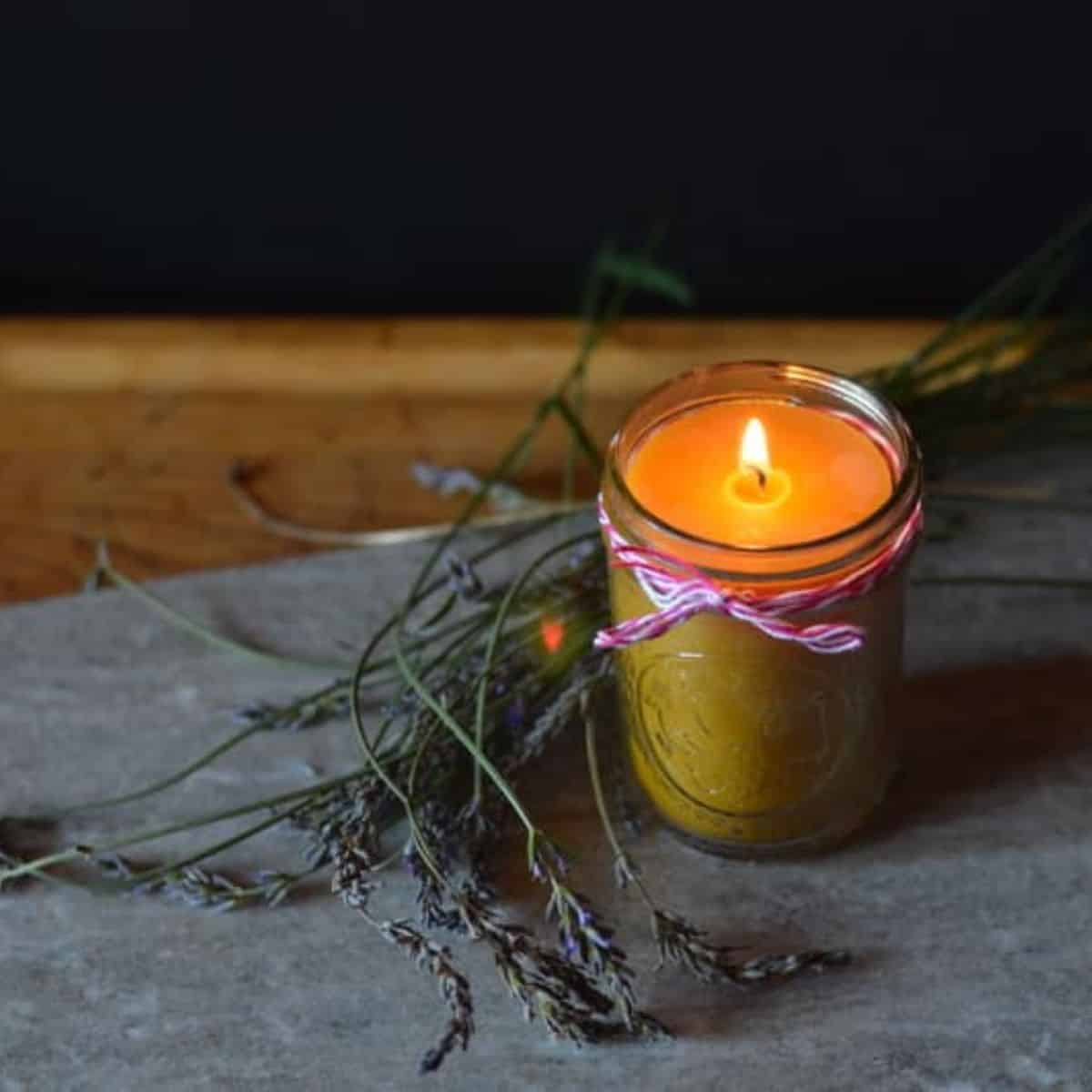 A lit beeswax candle in a glass jar, tied with pink and white string, sits on a stone surface next to sprigs of dried lavender against a dark background.