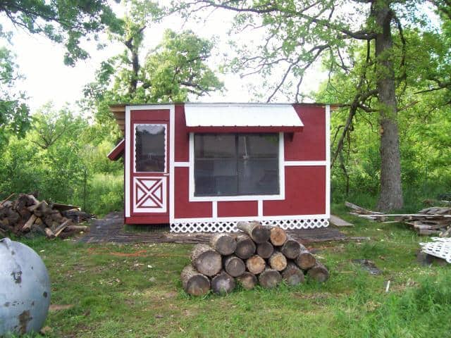 A small red and white shed with a metal roof stands in a grassy area surrounded by trees, ideal for transforming into a DIY chicken coop. Logs are stacked in front, and to the side, a propane tank and wooden planks await use. The scene is bright with natural daylight.