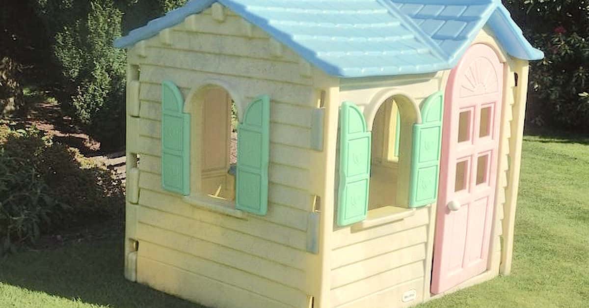 A small, plastic playhouse with a light blue roof and pastel walls stands on a grassy lawn, ingeniously repurposed as a DIY chicken coop. It features pink doors, green shutters, and arched windows, surrounded by greenery under bright sunlight.