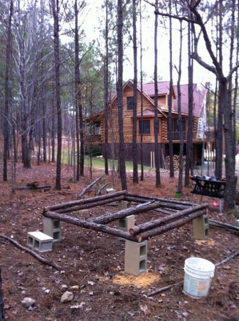 A wooden cabin surrounded by trees in a forest setting. In the foreground, there's a DIY chicken coop with a square frame made of wooden logs, supported by concrete blocks, and a white bucket on the ground.