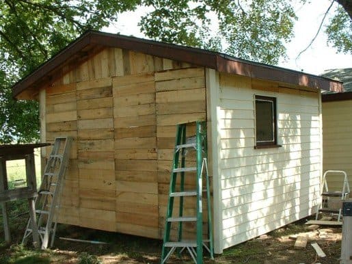 A small wooden shed, envisioned as a DIY chicken coop, is under construction. Unfinished wood planks adorn one side while white siding completes the other. Two ladders stand ready for use, framed by the gentle shadows of surrounding trees.