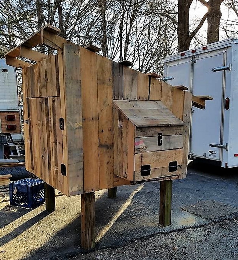 A DIY chicken coop crafted from repurposed planks stands elevated on four posts. It features a small entrance and a nesting box on the side. The structure is set outdoors, with bare trees and a parked trailer in the background.