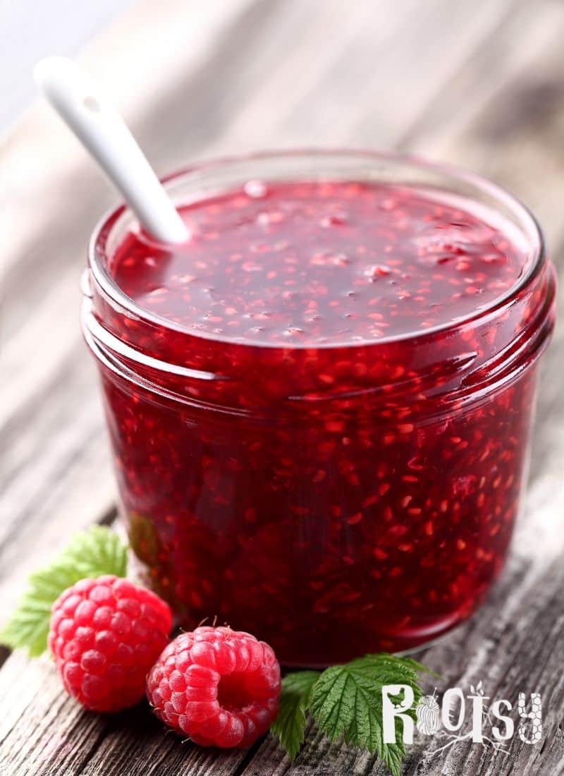 image of raspberry jam in canning jar with white spoon and two raspberries on wooden table