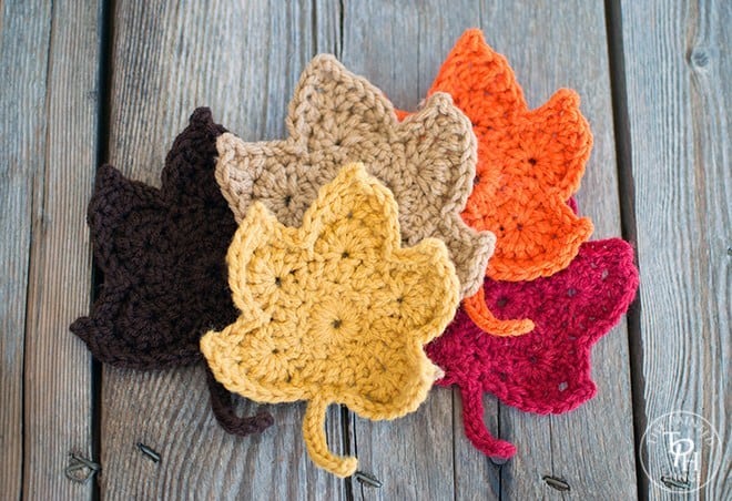 A collection of crocheted leaves in rich autumn colors—brown, beige, orange, yellow, and red—arranged artfully on a wooden surface embodies the essence of fall crafts.