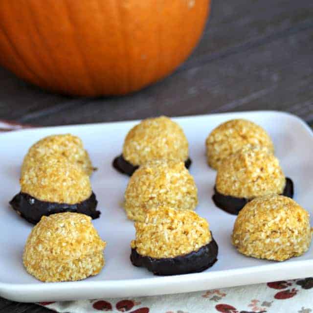 A plate of dessert balls with orange-colored tops, hinting at pumpkin puree, and dark chocolate-dipped bottoms, neatly arranged on a white rectangular plate. A pumpkin in the background sits on a wooden table, inspiring autumnal dessert recipes.