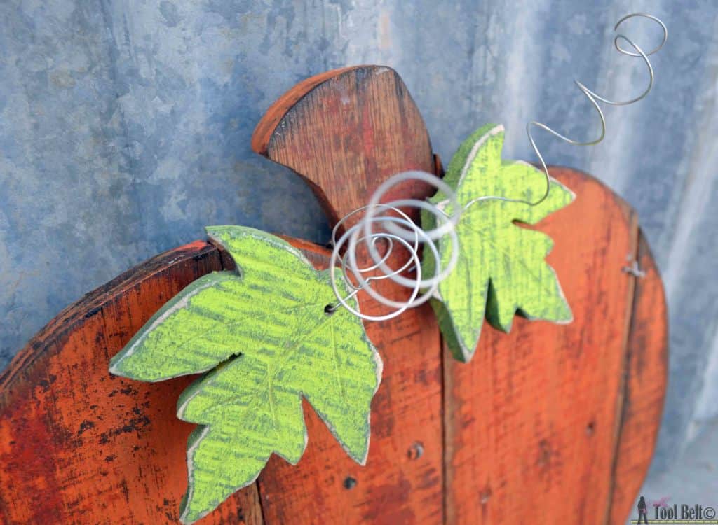 Wooden pumpkin decoration with two painted green leaves and a coiled metal wire resembling a vine, set against a corrugated metal background.