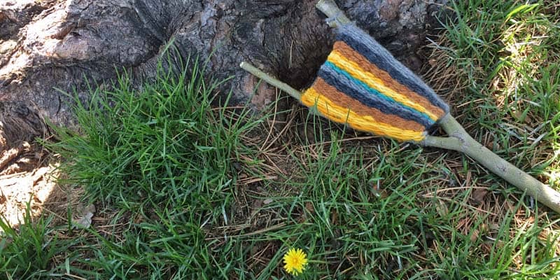 A small tree branch wrapped with colorful yarn lies on green grass beside a tree trunk. A single yellow dandelion blooms nearby, adding a touch of charm to this fall crafts scene.