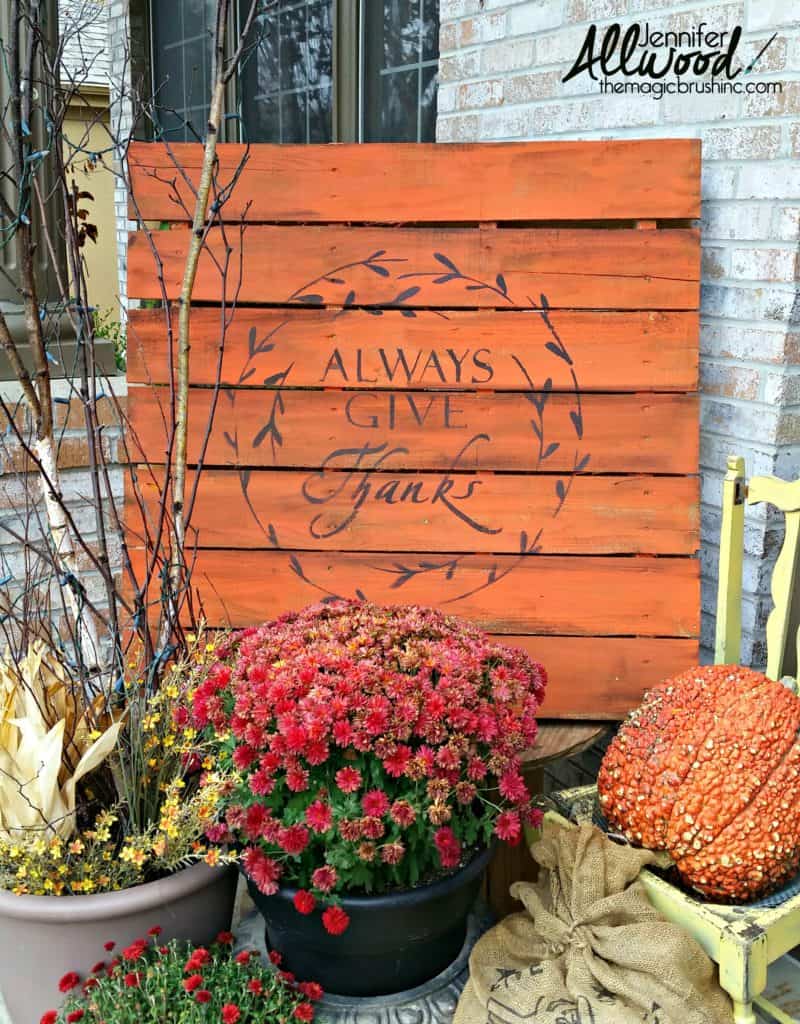 Wooden sign with "Always Give Thanks" written on it, surrounded by potted autumn flowers and a large pumpkin. Brick wall backdrop.