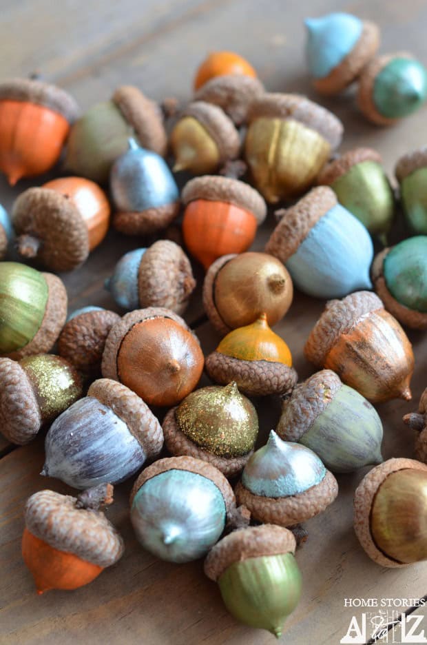 A collection of acorns painted in vibrant metallic colors, including silver, gold, blue, green, and orange, scattered on a wooden surface. This dazzling display of fall crafts transforms simple acorns into shimmering treasures.