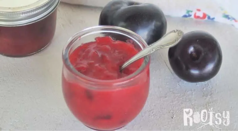 An open jar of plum jam with spoon inside it on a table with fresh plums and cloth napkin. 