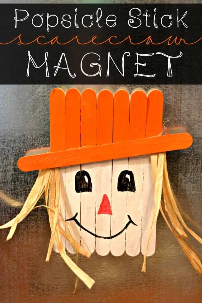 A popsicle stick scarecrow magnet with a smiling face, orange hat, and straw hair is displayed against a metallic background. Playful lettering above reads "Popsicle Stick Scarecrow Magnet." Perfect for fall crafts, this charming creation adds a touch of seasonal joy to any space.