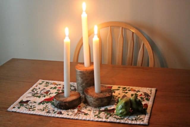 Three lit white candles rest on rustic wood bases atop a patterned table runner, complemented by a green ceramic artichoke. This inviting scene, perfect for fall crafts inspiration, features a wooden chair poised at the wooden table in the background.