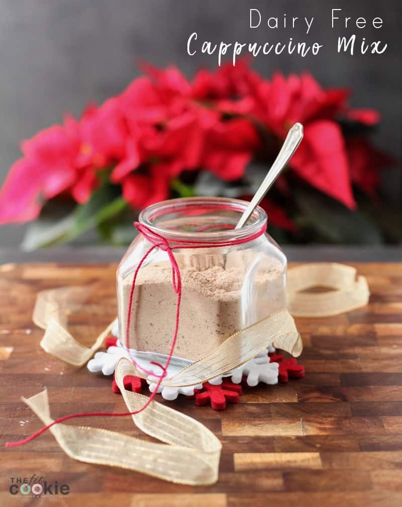 A jar of dairy-free cappuccino mix with a spoon, adorned with a red ribbon, sits invitingly on a wooden surface. Red poinsettias in the background add a festive touch, making it perfect for gifts in a jar. The text "Dairy Free Cappuccino Mix" graces the top right corner.