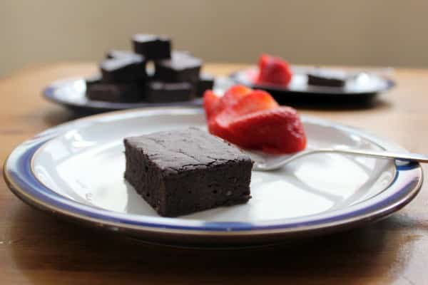 A slice of dark chocolate cake on a white plate, accompanied by sliced strawberries. Another plate with more cake pieces and strawberries is visible in the background on a wooden table.