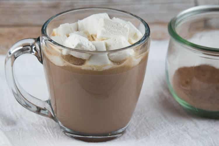 A glass mug of hot chocolate heaped with marshmallows sits invitingly, while a small jar filled with cocoa powder—a perfect gifts in a jar idea—rests on a white cloth. The cozy scene unfolds on a rustic wooden surface, capturing the essence of warmth and indulgence.