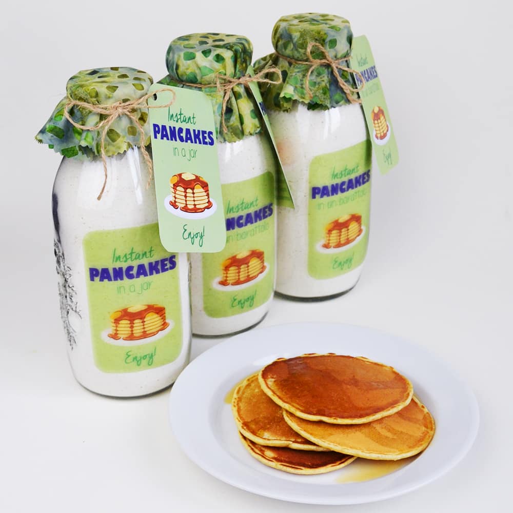 Three glass jars with labeled tags containing instant pancake mix are displayed behind a white plate with a stack of golden-brown pancakes. The jars, perfect as gifts in a jar, have decorative covers and tags reading "Instant Pancakes in a Jar.