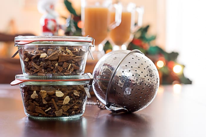 Two glass jars filled with dried spices hint at delightful gifts in a jar, sitting next to a large metal tea infuser. In the background, blurred festive decorations and glasses with an orange beverage create a cozy, celebratory atmosphere.