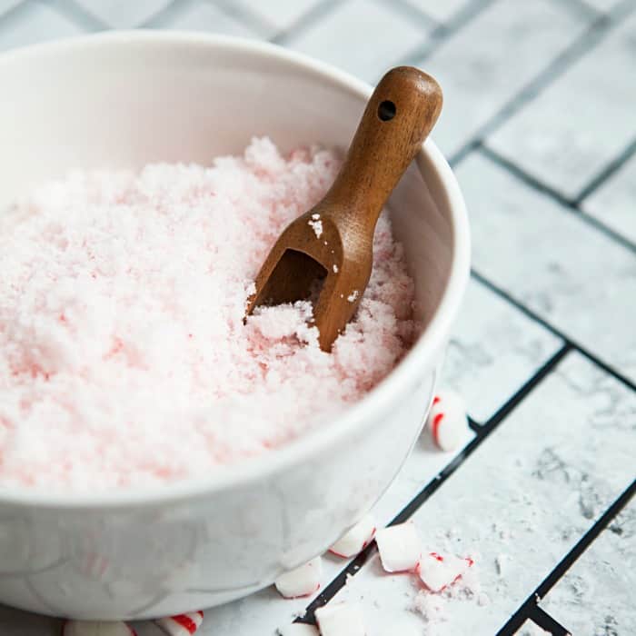 A white bowl filled with pink and white granular sugar, reminiscent of gifts in a jar, resembling coarse salt, with a wooden scoop partially buried in it. The bowl is elegantly placed on a tiled surface.