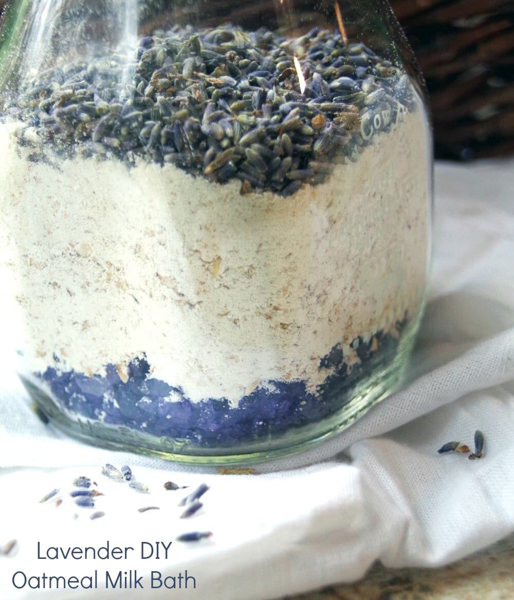 A glass jar layered with dried lavender, oatmeal, and a mysterious purple substance sits delicately on a white cloth, surrounded by sprigs of lavender. Text reads: "Lavender DIY Oatmeal Milk Bath." Perfect for thoughtful gifts in a jar.