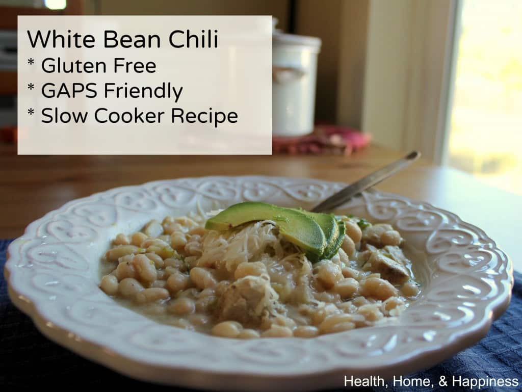 A bowl of white bean chili garnished with sliced avocado sits on a table. Text on the image says: "White Bean Chili * Gluten Free * GAPS Friendly * Slow Cooker Recipe." The corner text reads, "Health, Home, & Happiness.