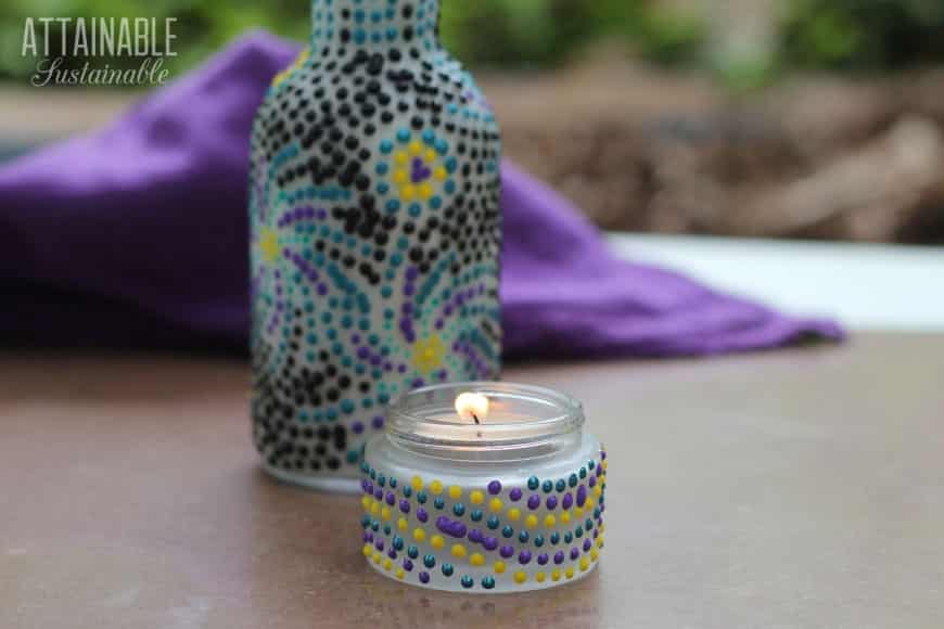 A small glass jar with a lit tealight candle, perfect for gifts in a jar, is decorated with colorful dots. Behind it stands a bottle adorned with a similar pattern. A purple cloth is partially visible in the background.