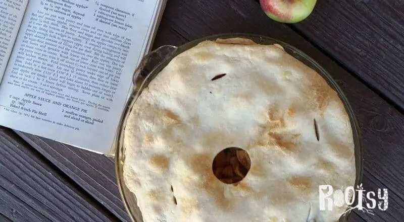A baked apple pie sitting next to a book and fresh apple.