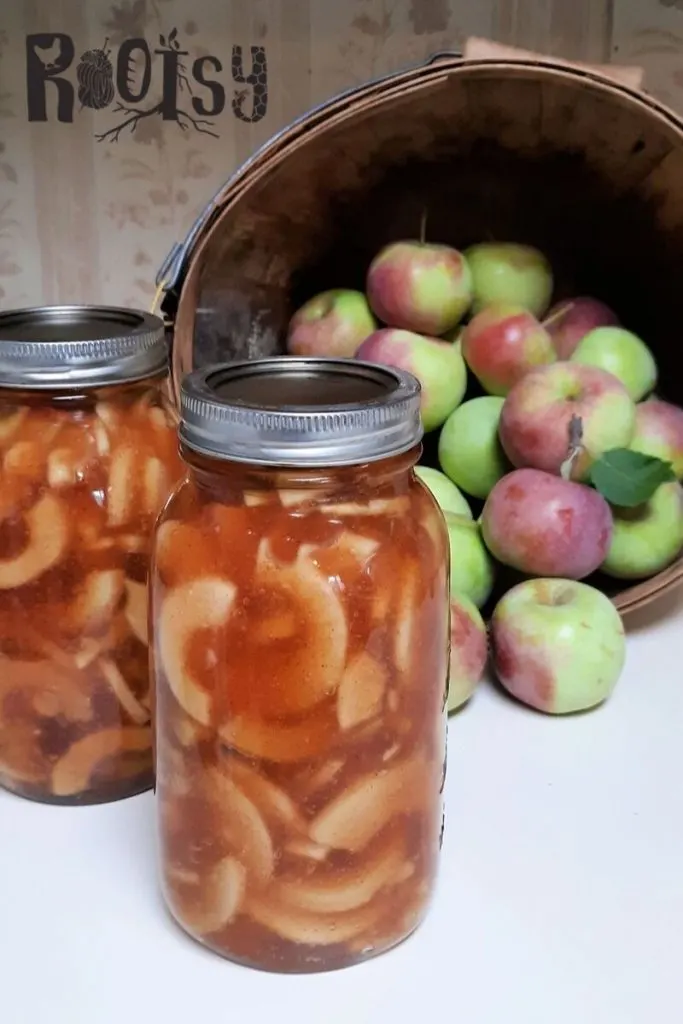 Jars of canned apple pie filling in front of a basket of fresh apples.