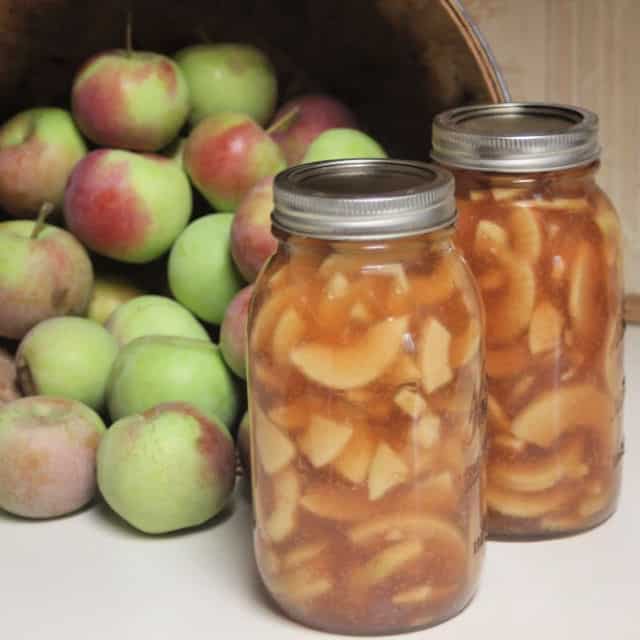 2 jars of canned apple pie filling sitting in front of a basket of fresh apples