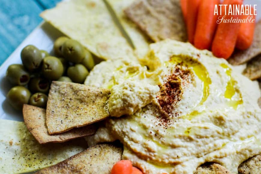 A plate of hummus topped with spices and olive oil is surrounded by olives, pita chips, and baby carrots. The background features a logo with the words "Attainable Sustainable.