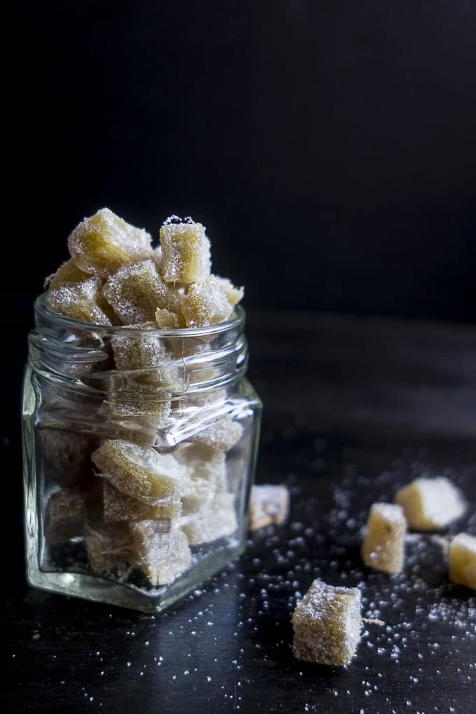 A glass jar filled with sugar-coated ginger pieces is placed on a dark surface, resembling delightful gifts in a jar. A few ginger pieces are scattered around, and the surface is sprinkled with sugar. The background is dark, highlighting the texture and sparkle of this sweet treat.