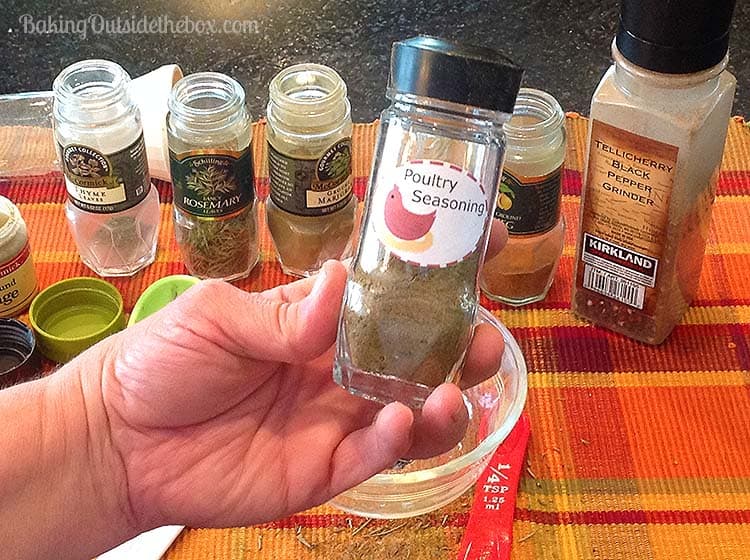 A hand holds a clear spice jar labeled "Poultry Seasoning" over a kitchen counter, resembling gifts in a jar. Various other spice jars, including rosemary and marjoram, grace the background, while a red measuring spoon rests beside a small bowl.