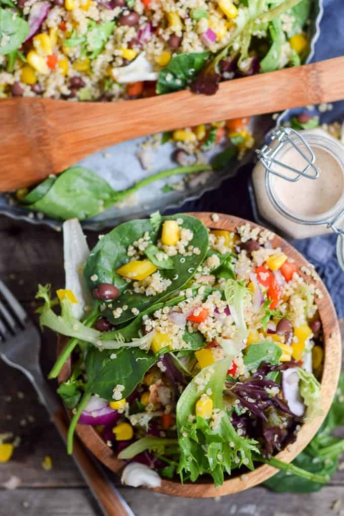 A wooden bowl filled with a colorful salad containing quinoa, mixed greens, black beans, corn, and diced red peppers. A dressing jar and a fork are beside it, with a larger bowl of salad in the background.