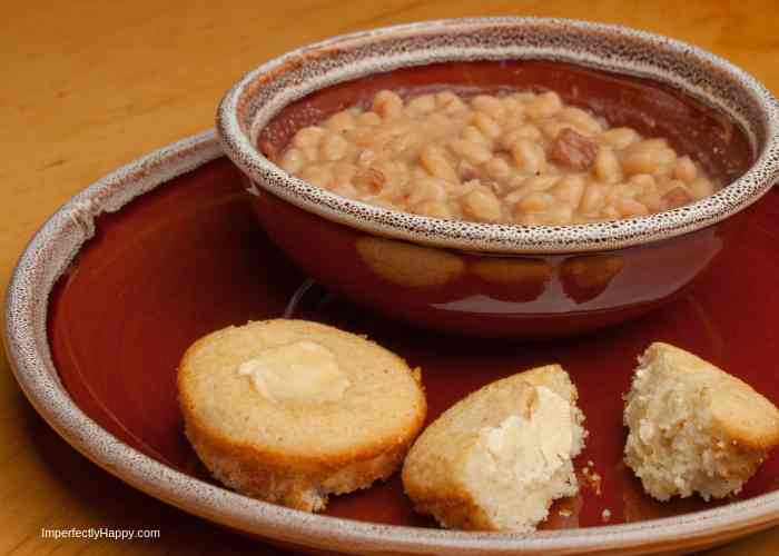 A ceramic bowl filled with baked beans sits on a matching plate. Next to the bowl, a cornbread muffin is broken into two pieces, with one piece topped with butter. The background is a wooden table.