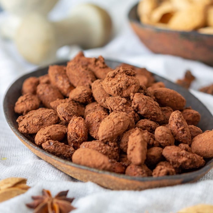 A close-up of a bowl filled with chocolate-coated almonds on a white textured cloth, perfect for gifts in a jar. In the background, there's a blurred bowl with other snacks. Star anise and dried leaves are scattered around for decoration.
