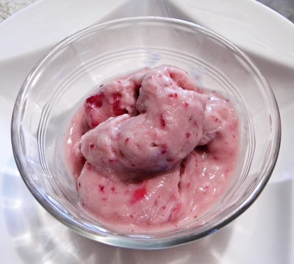 A glass bowl filled with creamy pink ice cream featuring visible chunks of strawberry, set on a white plate.