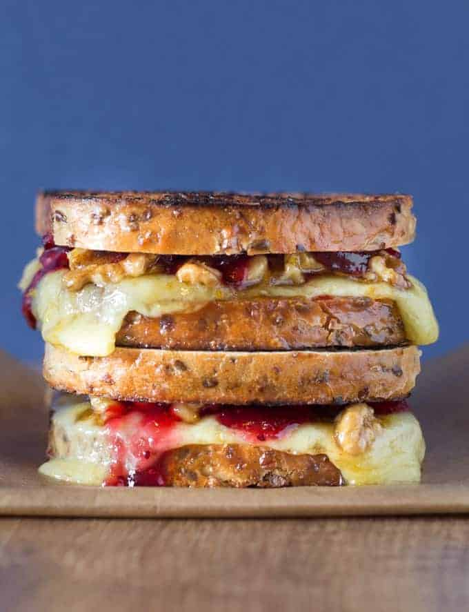 A close-up of a stacked grilled cheese sandwich with melted cheese, walnuts, and a red jam-like spread, on rustic multigrain bread, against a blue background.