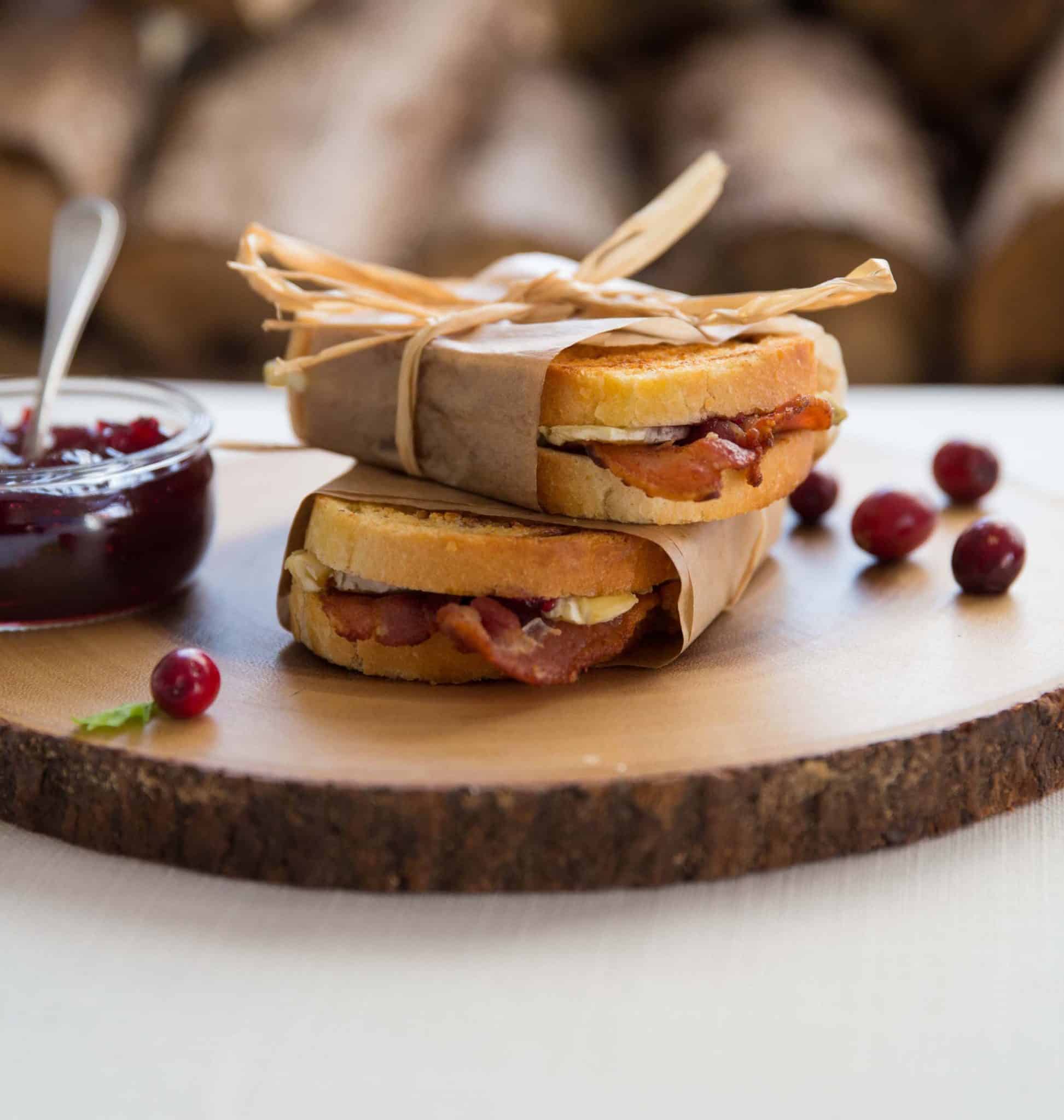 Two sandwiches wrapped in brown paper are placed on a wooden board. They're filled with bacon and cheese. A small jar of cranberry sauce is beside them, surrounded by a few fresh cranberries. Logs are visible in the blurred background.