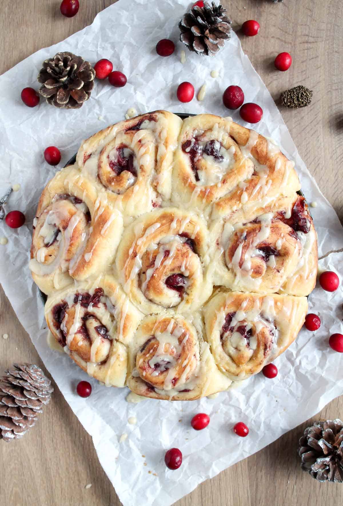 A tray of freshly baked cinnamon rolls with icing, arranged in a circle on parchment paper. Surrounded by red cranberries and pine cones on a wooden surface, creating a festive atmosphere.