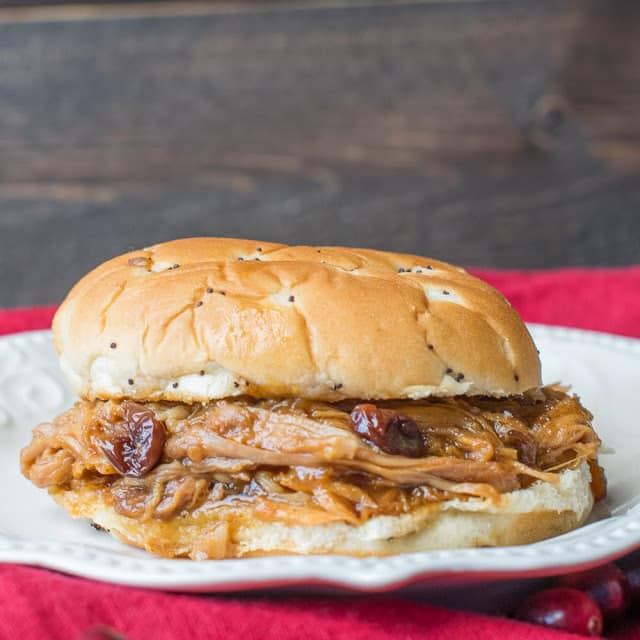 A pulled pork sandwich with cranberry sauce on a white plate. The sandwich is on a bun with poppy seeds, and the plate sits on a red cloth. The background is a dark wooden surface.