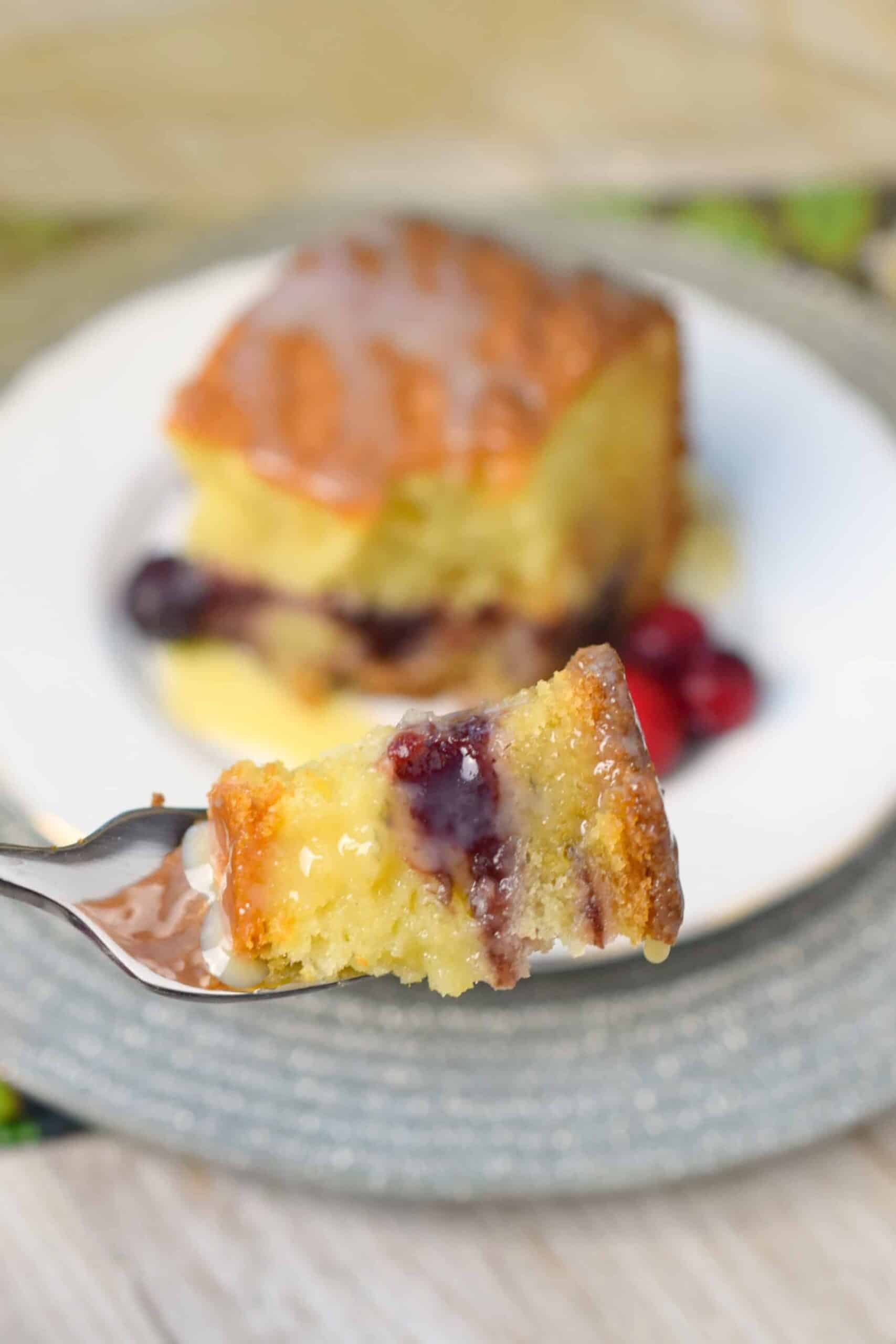 A close-up of a fork holding a piece of moist cake with layers of yellow cake and a red berry filling. The background shows a larger piece of the cake on a white plate, drizzled with glaze. Berry fruits are visible beside the cake.