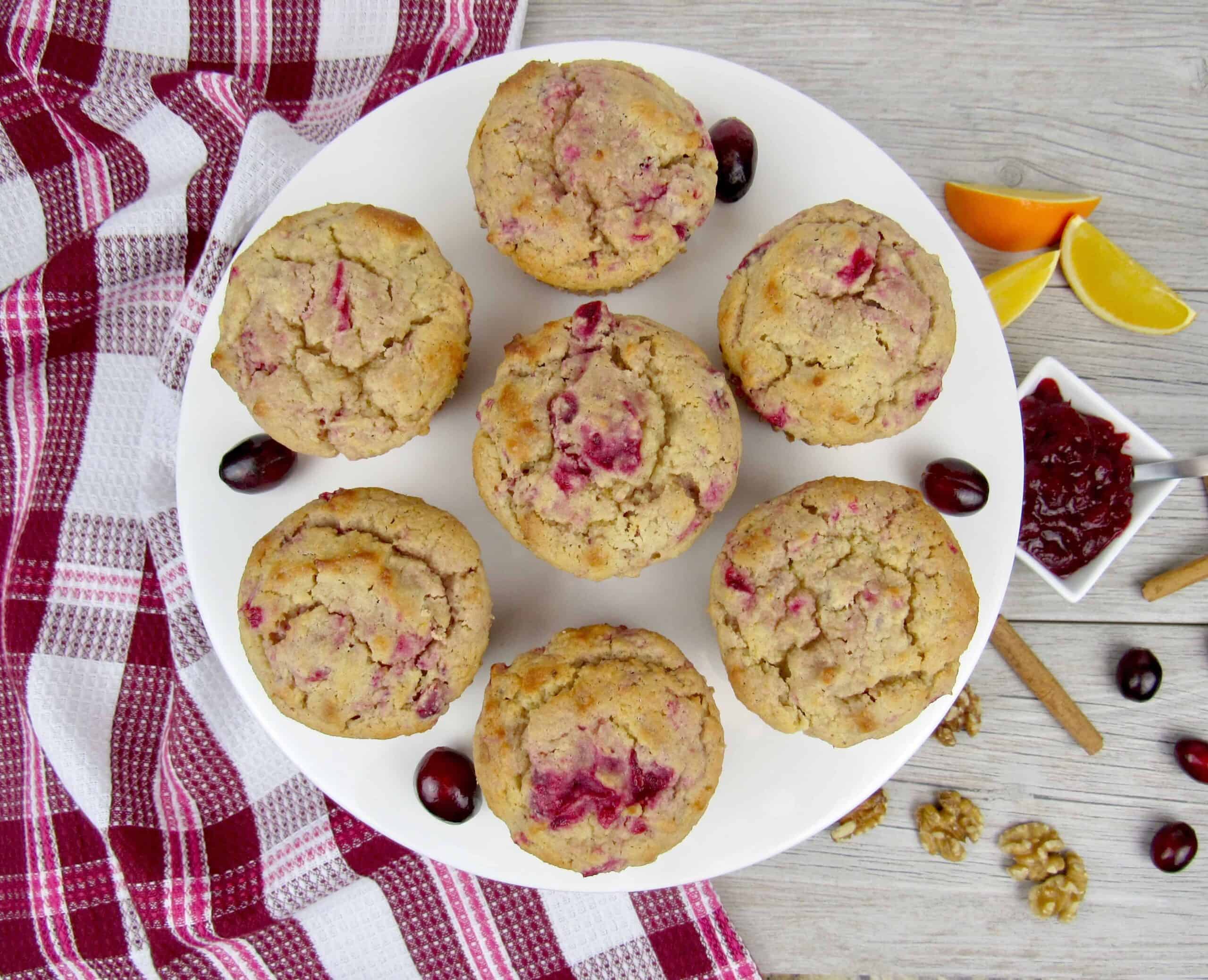 A white plate holds seven cranberry muffins on a wooden surface. A red and white checkered cloth is on the left. There are scattered cranberries, walnuts, orange slices, and a small dish of jam on the right.
