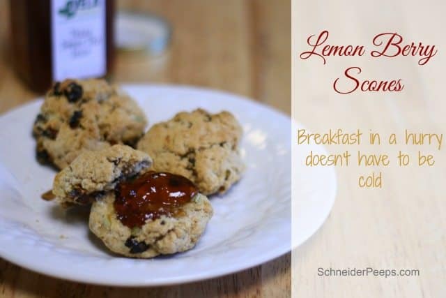 A plate of lemon berry scones, crafted from a premium baking mix, with a circular dollop of jam on top, rests on a wooden table. Text on the image reads: "Lemon Berry Scones: Breakfast in a hurry doesn't have to be cold." A jar in the background is slightly out of focus.