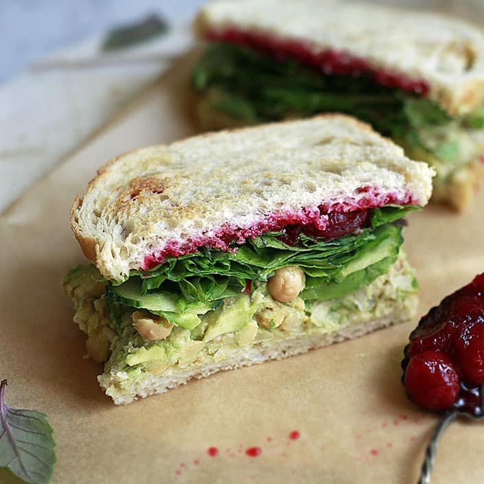 A sandwich with mashed avocado, chickpeas, leafy greens, and cranberry sauce on whole grain bread, placed on parchment paper. A spoonful of cranberry sauce is beside the sandwich. Another sandwich is partially visible in the background.